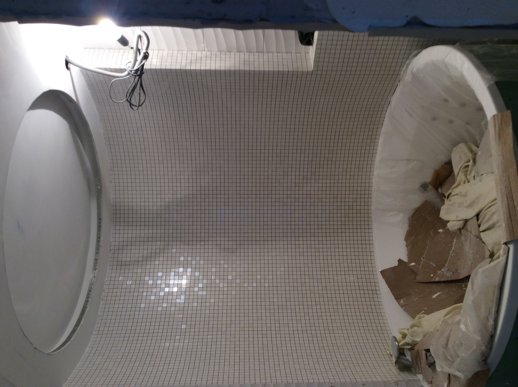 Wnętrze łazienki w trakcie remontu, widok z góry na zaokrągloną ścianę i podłogę wyłożoną drobną, jasną mozaiką. Wanna zabezpieczona folią i wypełniona odpadami budowlanymi. Widoczne przewody...