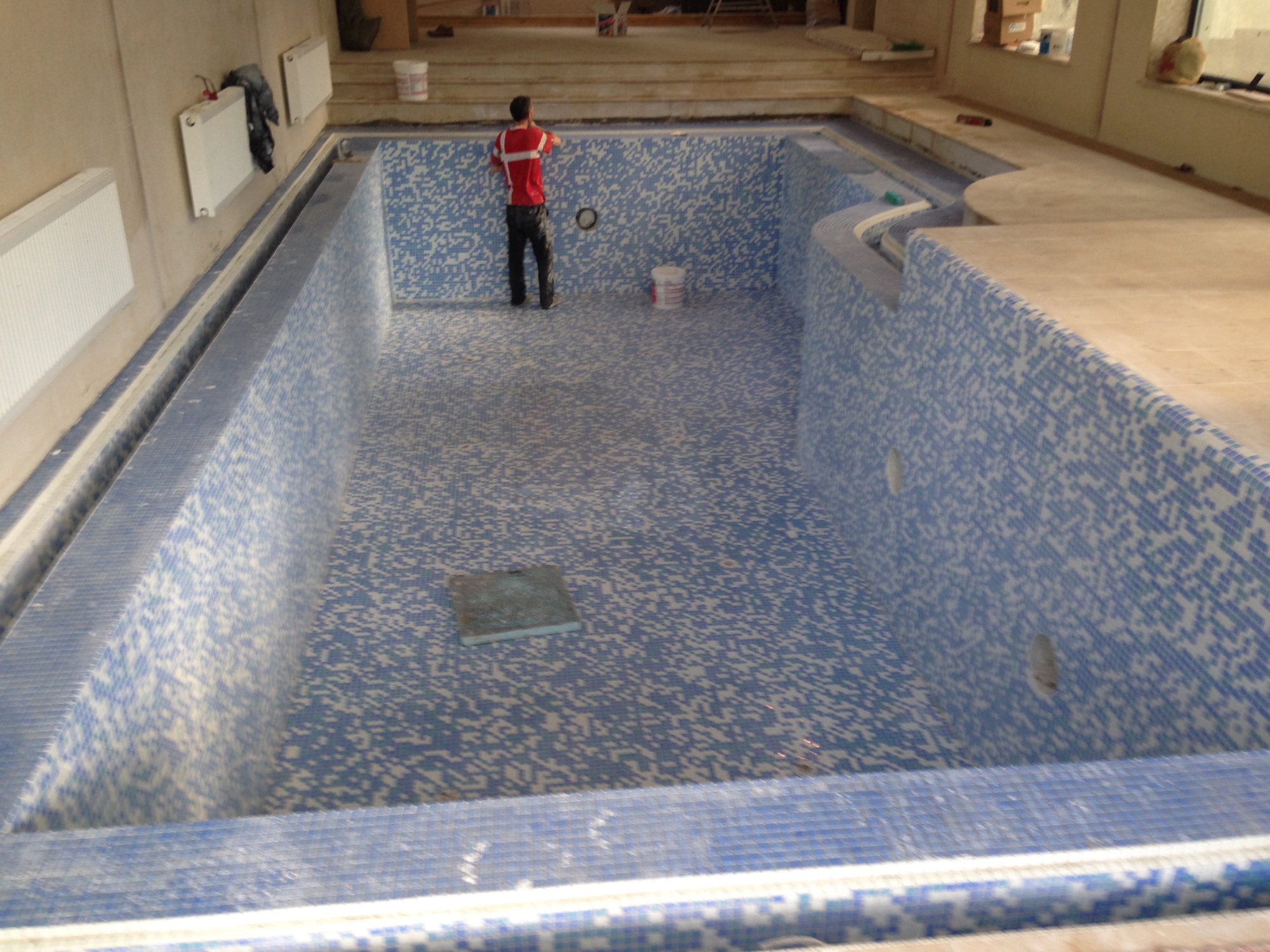 Wnętrze basenu w trakcie wykańczania, z mozaiką w odcieniach błękitu i białego, widoczny pracownik w czerwonej koszulce i czarnych spodniach przy ścianie basenu oraz wiadro z zaprawą.