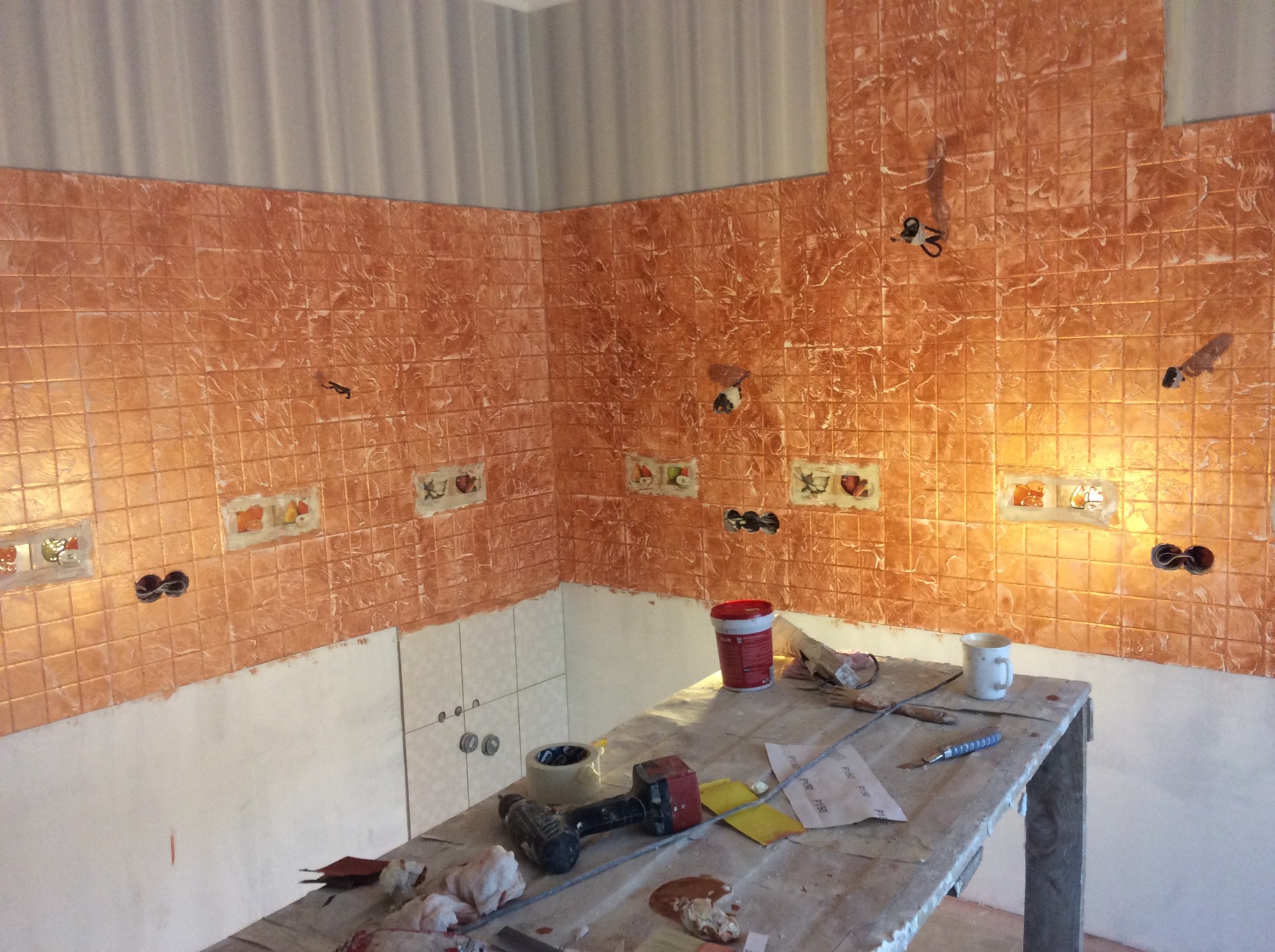 Kuchnia w trakcie remontu: pomarańczowe płytki imitujące kamień z dekoracyjnymi wstawkami, częściowo położone, widoczne puszki elektryczne, przygotowane narzędzia i materiały na stole roboczym.