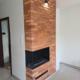 Nowoczesny kominek z cegły klinkierowej z czarnym wkładem, oświetlony punktowym światłem, w surowym, jasnym wnętrzu.