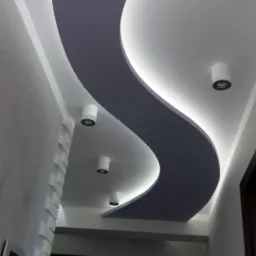 Sufit z płyt karton gips z podświetleniem LED w kształcie litery S, oświetlenie punktowe, dekoracyjna ściana z białych elementów, widok z dołu.