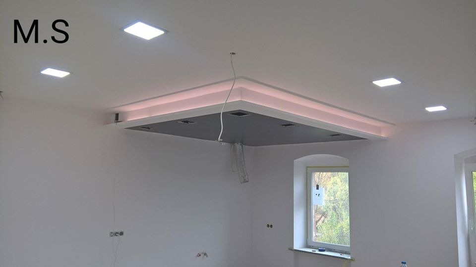 Podwieszany sufit z oświetleniem LED w trakcie montażu, widoczne przewody i elementy konstrukcyjne, białe ściany i okna w tle.