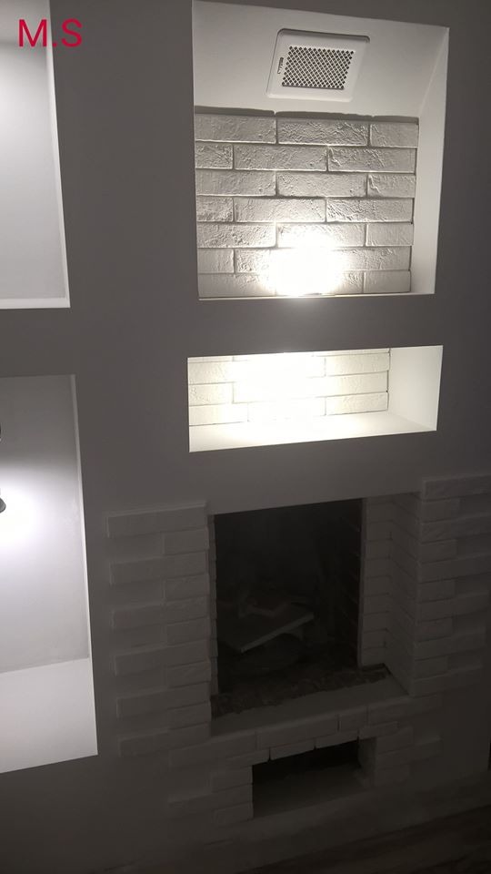 Nowoczesna ściana z wnękami, wykończona białą cegłą dekoracyjną, z podświetleniem LED w górnej wnęce i imitacją kominka na dole.