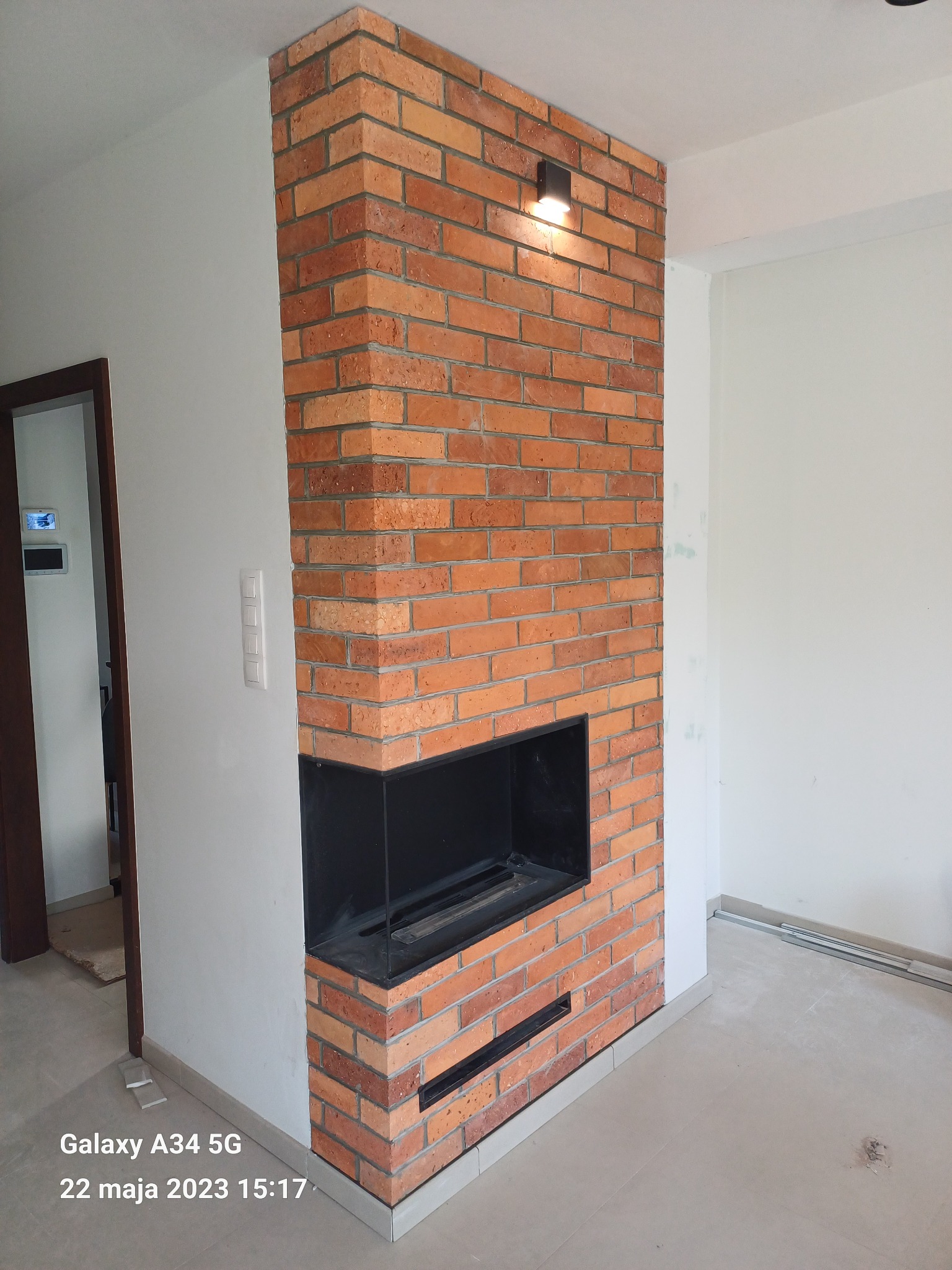 Nowoczesny kominek z cegły klinkierowej z czarnym wkładem, oświetlony punktowym światłem, w surowym, jasnym wnętrzu.