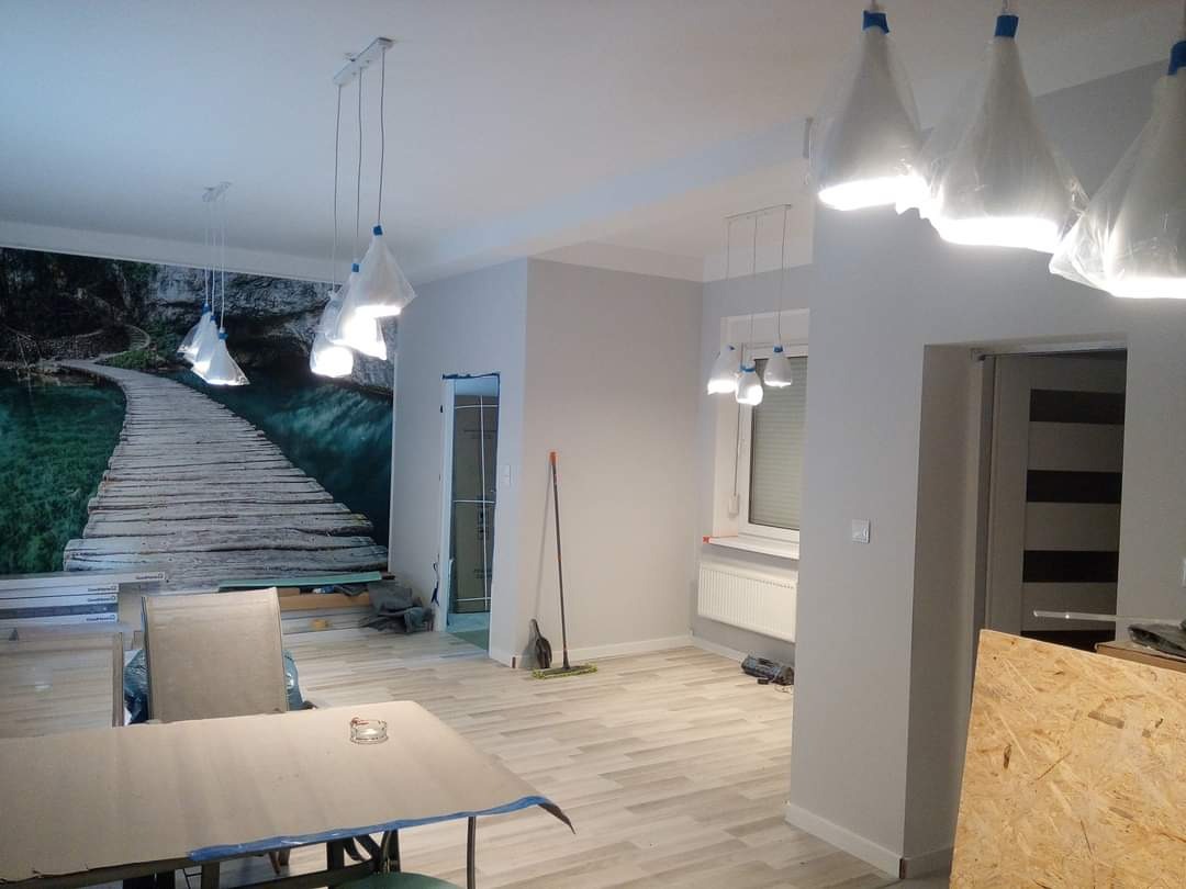 Wnętrze mieszkania w trakcie odświeżania: ściany pomalowane na szaro, na jednej ze ścian fototapeta z motywem drewnianego pomostu nad wodą, lampy wiszące zabezpieczone folią, podłoga wyłożona...