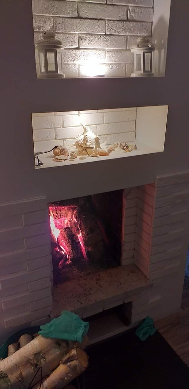 Oświetlony kominek z obudową z białej cegły, nad nim półka z dekoracjami morskimi, a na górze dwie latarenki.