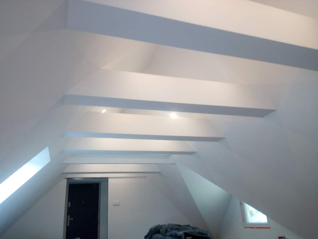 Wykończone poddasze ze skośnym sufitem i widocznymi belkami konstrukcyjnymi, pomalowane na biało, z oknem dachowym i drzwiami po lewej stronie.