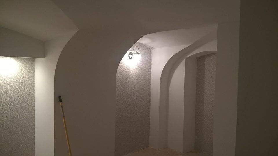 Wnętrze z łukowatymi przejściami pomalowane na biało i szaro, z widoczną lampą ścienną i fragmentem narzędzia malarskiego.