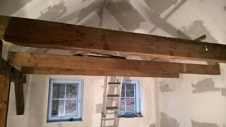 Wnętrze remontowanego pomieszczenia z widocznymi drewnianymi belkami stropowymi, fragmentami płyt na suficie, aluminiową drabiną oraz dwoma oknami z niebieskimi ramami.