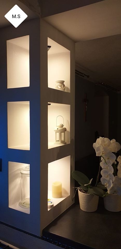 Ściana z wnękami oświetlonymi punktowo, w których umieszczono dekoracje: świecę, lampion, szklany pojemnik i ceramiczną figurkę, obok storczyki w doniczkach.