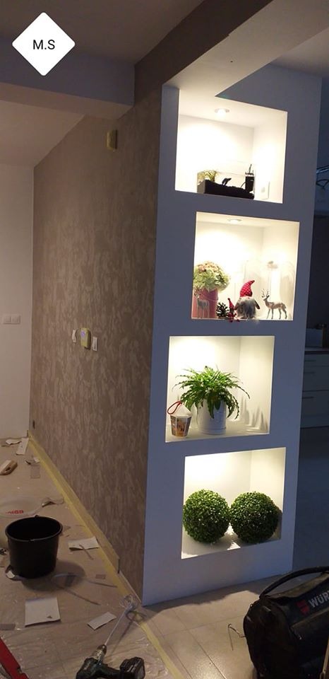 Ściana z dekoracyjną fakturą i wnękami z oświetleniem, w których umieszczono ozdoby, rośliny i akwarium, widoczne narzędzia malarskie i zabezpieczenia podłogi.