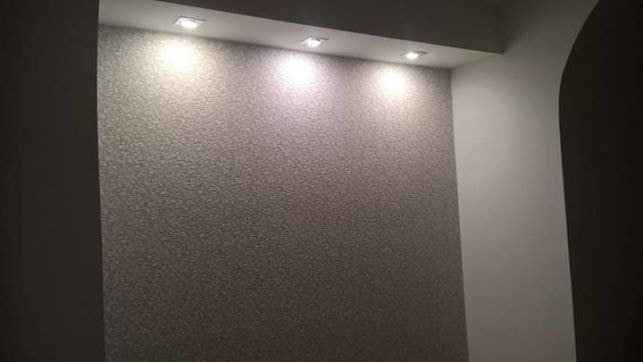 Teksturowana ściana w odcieniach szarości oświetlona trzema punktowymi lampami sufitowymi, widoczny fragment białej ściany po prawej stronie.