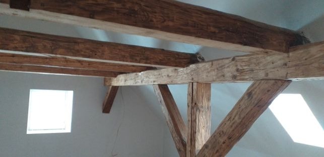 Wnętrze poddasza z drewnianą konstrukcją więźby dachowej, widoczne bielone ściany, okno dachowe i małe okienko na ścianie bocznej.