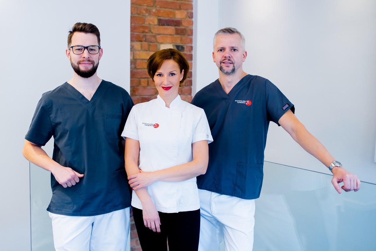 Zespół stomatologiczny: dwoje lekarzy w granatowych uniformach i lekarka w białym fartuchu na tle białej ściany i fragmentu ściany z czerwonej cegły.