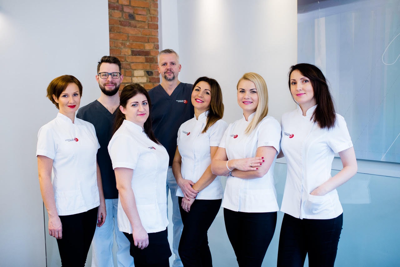 Zespół stomatologiczny w białych uniformach z logo kliniki, pozujący w jasnym wnętrzu z ceglaną ścianą w tle.