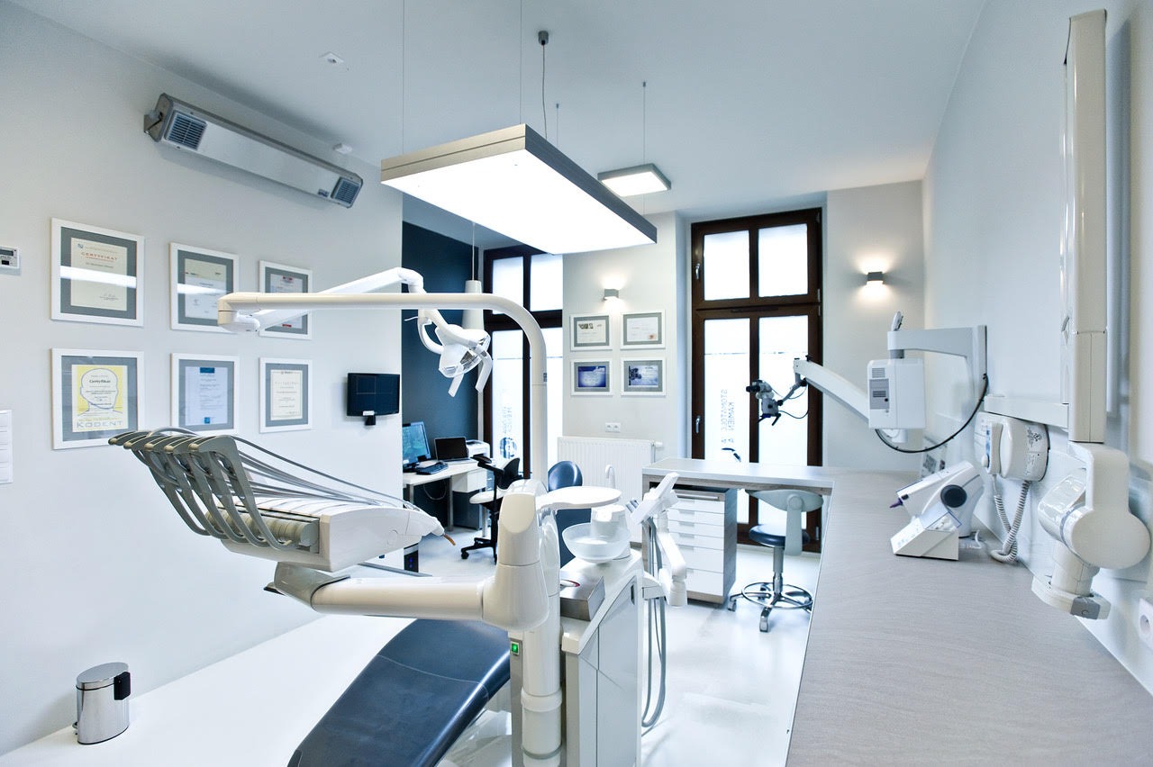 Nowoczesny gabinet lekarski z fotelem dentystycznym, oświetleniem LED, biurkiem z komputerem i ścianą z certyfikatami w ramkach.