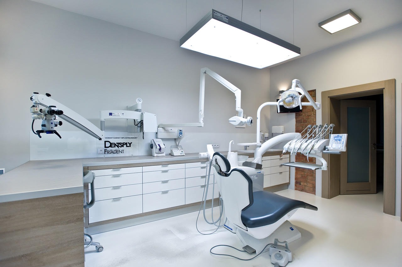 Jasne, przestronne wnętrze gabinetu stomatologicznego z nowoczesnym wyposażeniem, w tym mikroskopem zabiegowym i fotelem pacjenta z zestawem narzędzi.