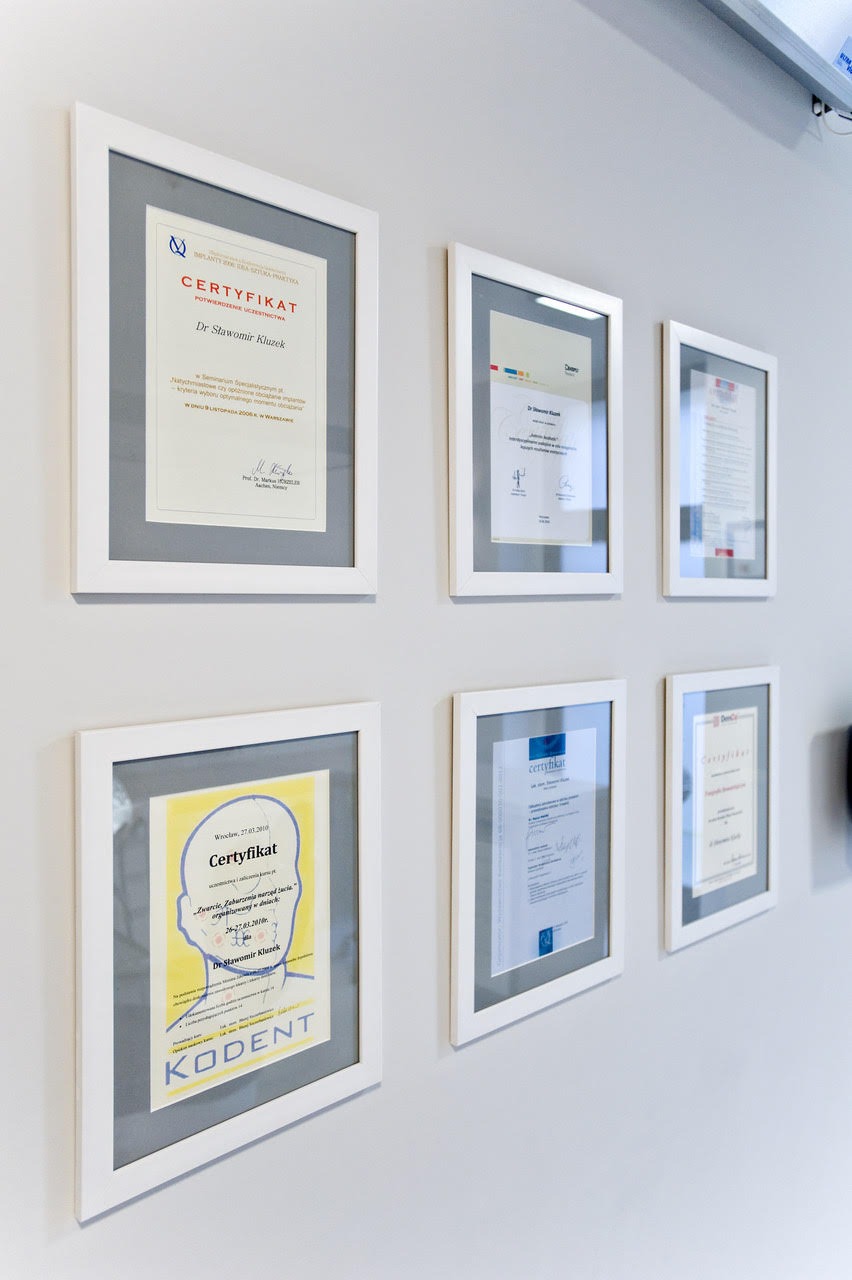 Ściana gabinetu z powieszonymi w białych ramkach certyfikatami i dyplomami lekarza specjalisty.