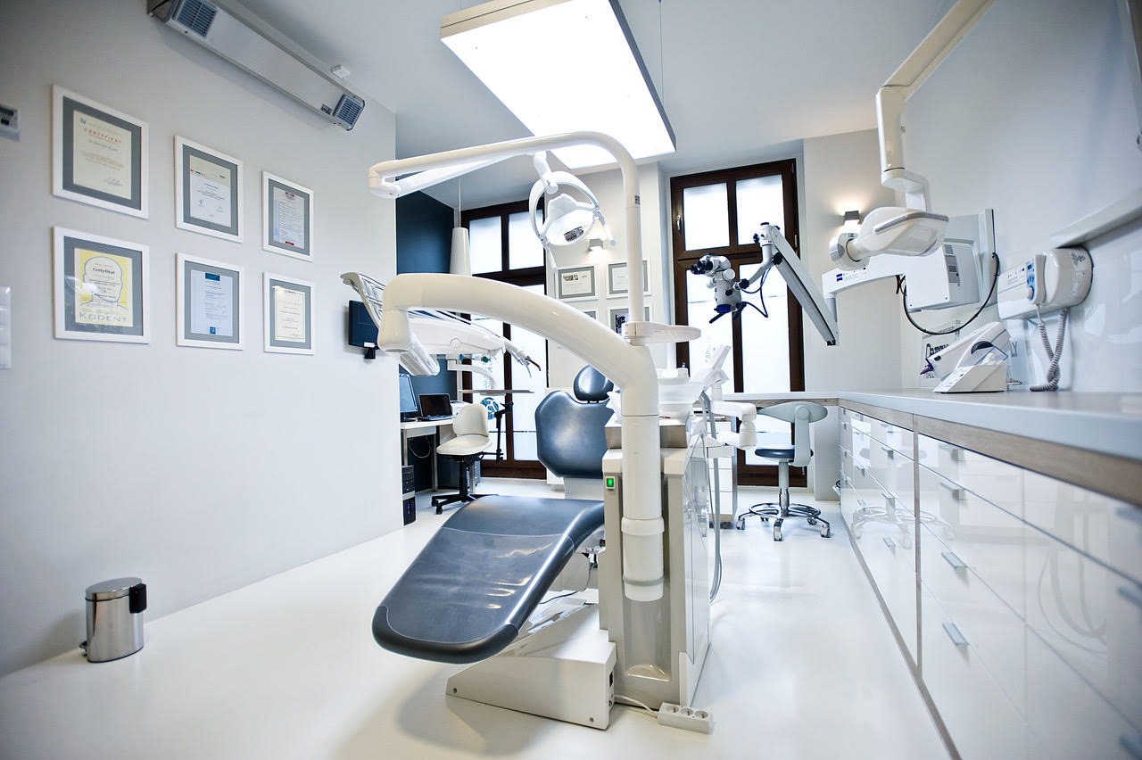 Jasne, sterylne wnętrze gabinetu lekarskiego z nowoczesnym fotelem stomatologicznym, mikroskopem zabiegowym i ścianą z certyfikatami w ramkach.