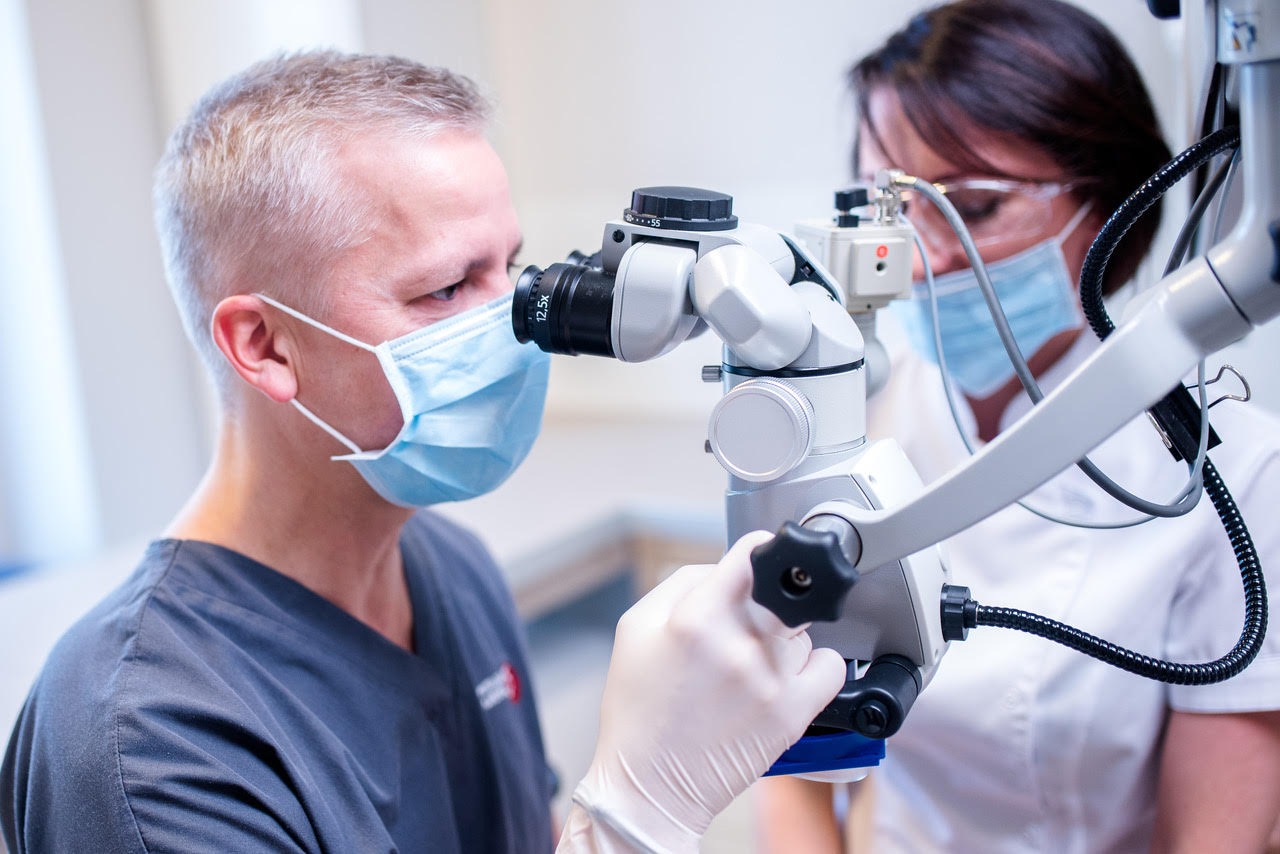 Lekarz w masce i rękawicach, pochylony nad mikroskopem zabiegowym, obok asystentka w okularach ochronnych i masce.