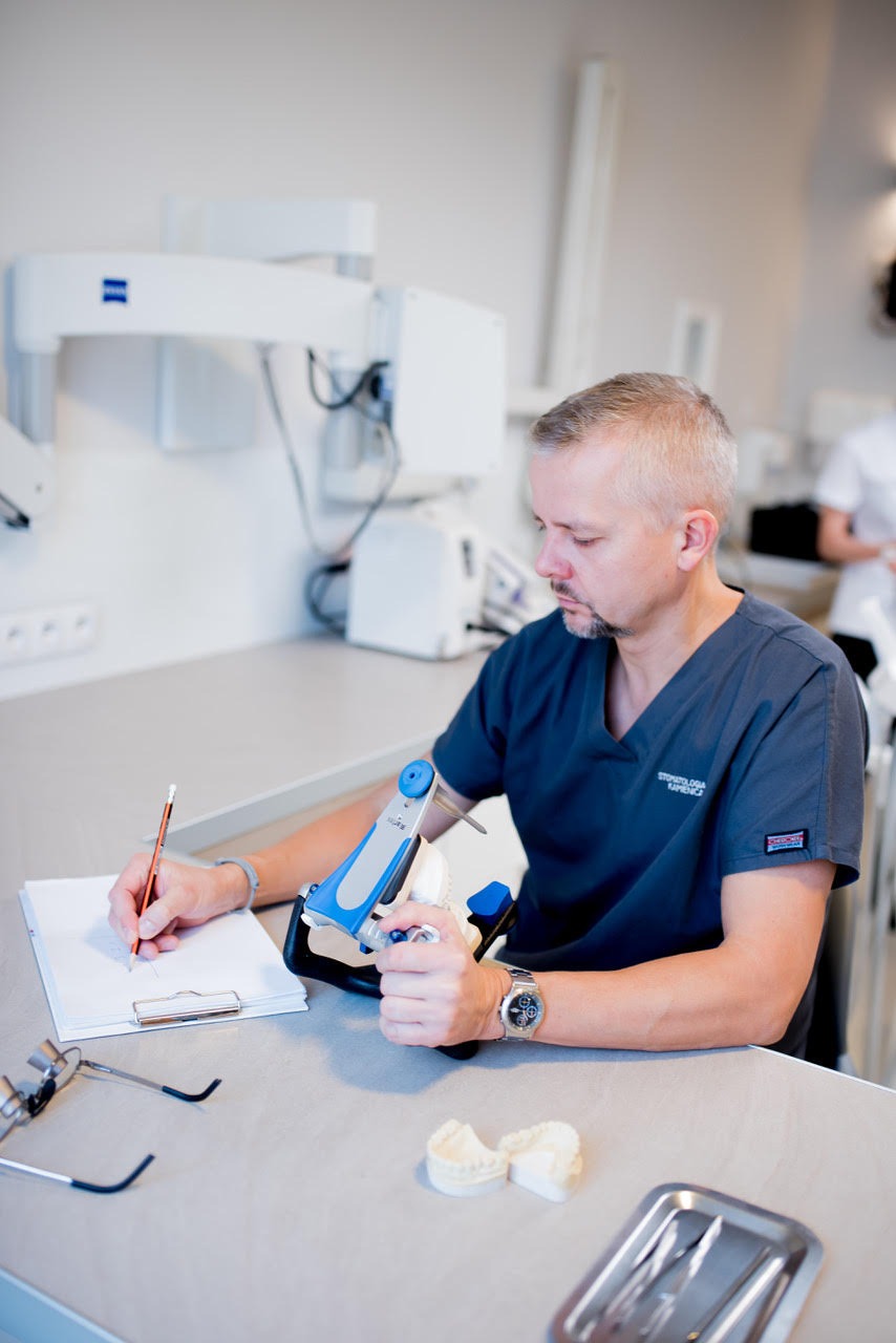 Stomatolog w ciemnoniebieskim uniformie z logo 'Stomatologia Kamienicz' zapisuje pomiary z artykulatora na kartce, obok model szczęki i narzędzia na tacy.