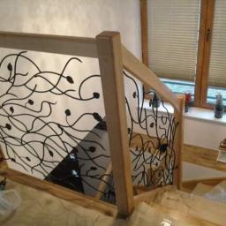 Kuźnia Ślusarstwo Spawanie - Drewniana balustrada schodowa z dekoracyjnym, metalowym wypełnieniem o motywie roślinnym, widok z góry na schody w trakcie montażu, narzędzia pracy obok.