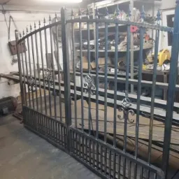 Metalowa brama dwuskrzydłowa z dekoracyjnymi elementami, w trakcie produkcji w warsztacie, widoczne narzędzia i materiały na półkach w tle.