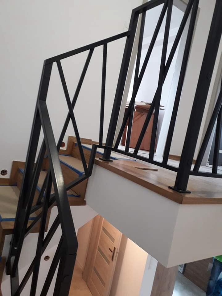 Nowoczesna, czarna, metalowa barierka schodowa z geometrycznym wzorem na drewnianych schodach w trakcie montażu, zabezpieczonych niebieską taśmą malarską.