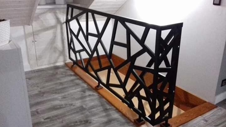 Czarna, metalowa balustrada schodowa o geometrycznym wzorze w nowoczesnym wnętrzu z szarymi panelami podłogowymi.