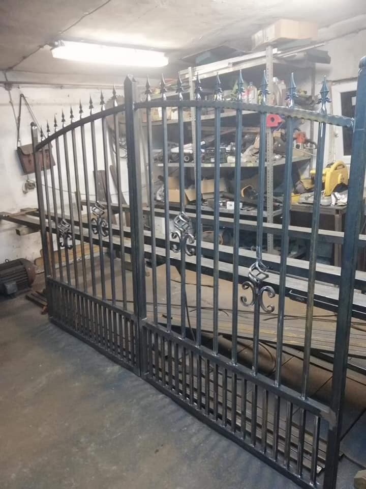 Metalowa brama dwuskrzydłowa z dekoracyjnymi elementami, w trakcie produkcji w warsztacie, widoczne narzędzia i materiały na półkach w tle.