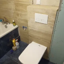 Nowoczesna toaleta podwieszana z białym przyciskiem spłukującym Kolo, obudowana płytkami imitującymi drewno; widoczne również fragmenty wanny, niebieskich płytek podłogowych i niezakończonej ściany...