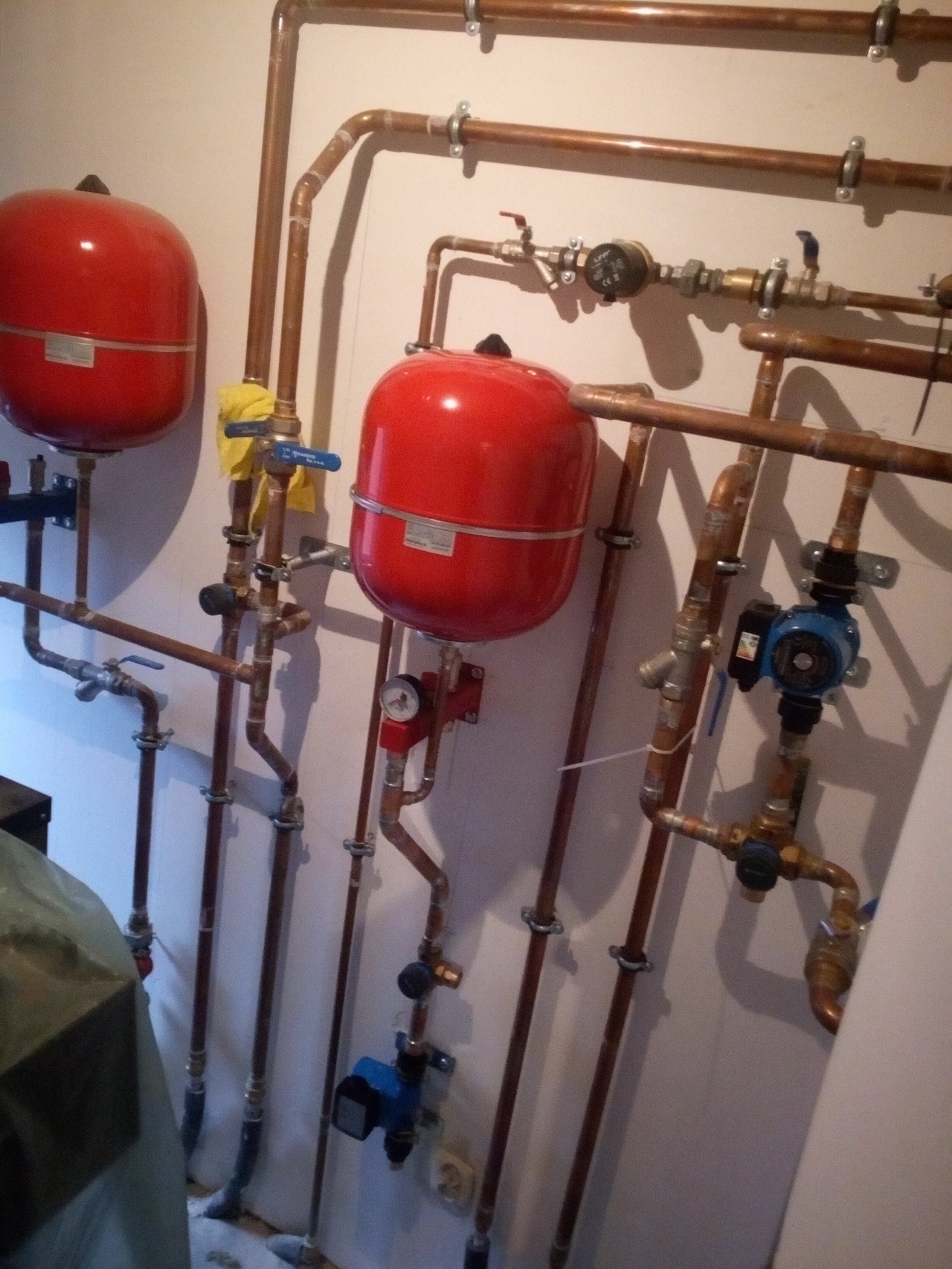 Kompleksowa instalacja centralnego ogrzewania z dwoma czerwonymi zbiornikami wyrównawczymi, licznymi miedzianymi rurami, zaworami i pompą obiegową, zamontowana na jasnej ścianie.