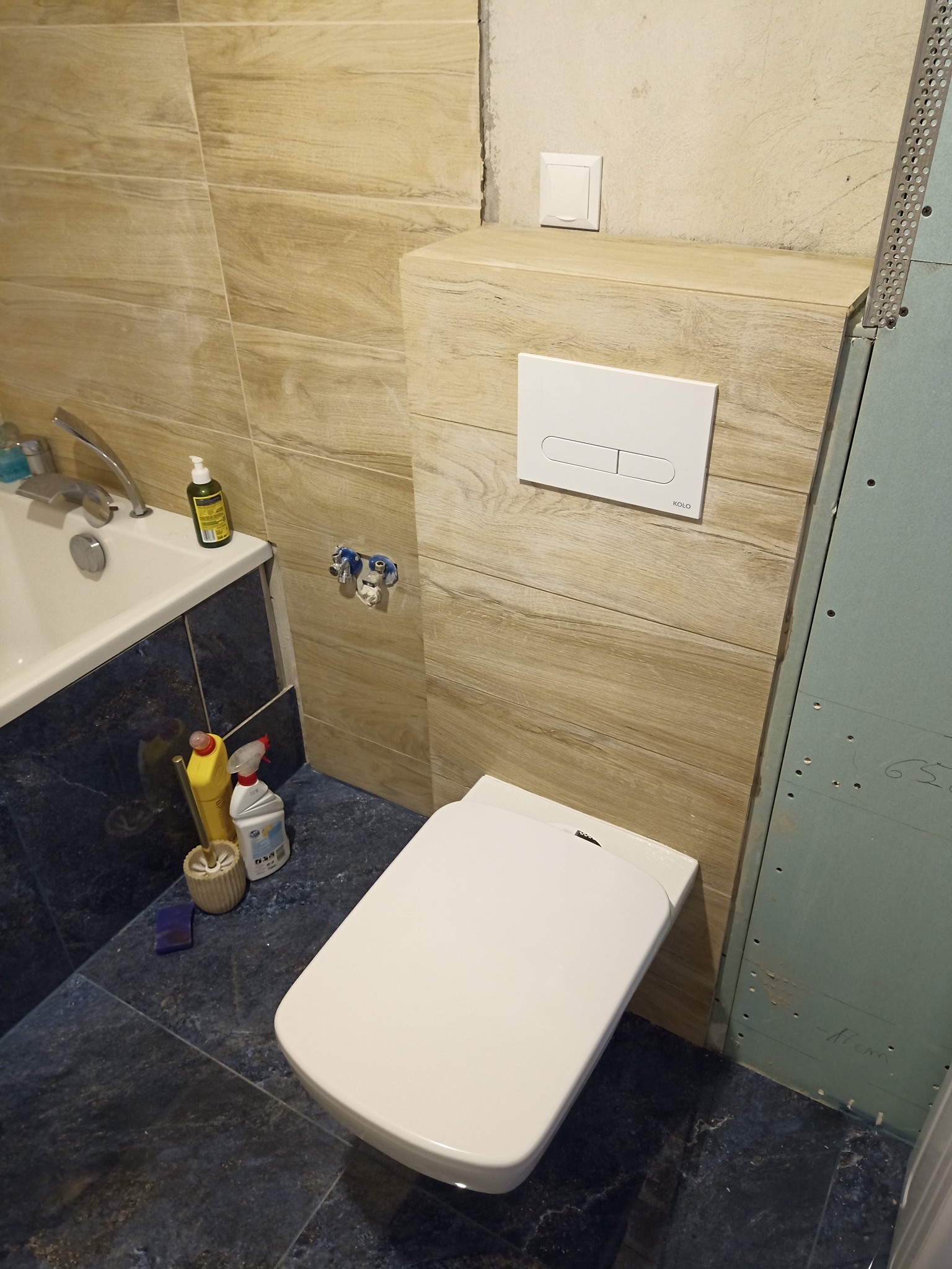 Nowoczesna toaleta podwieszana z białym przyciskiem spłukującym Kolo, obudowana płytkami imitującymi drewno; widoczne również fragmenty wanny, niebieskich płytek podłogowych i niezakończonej ściany...