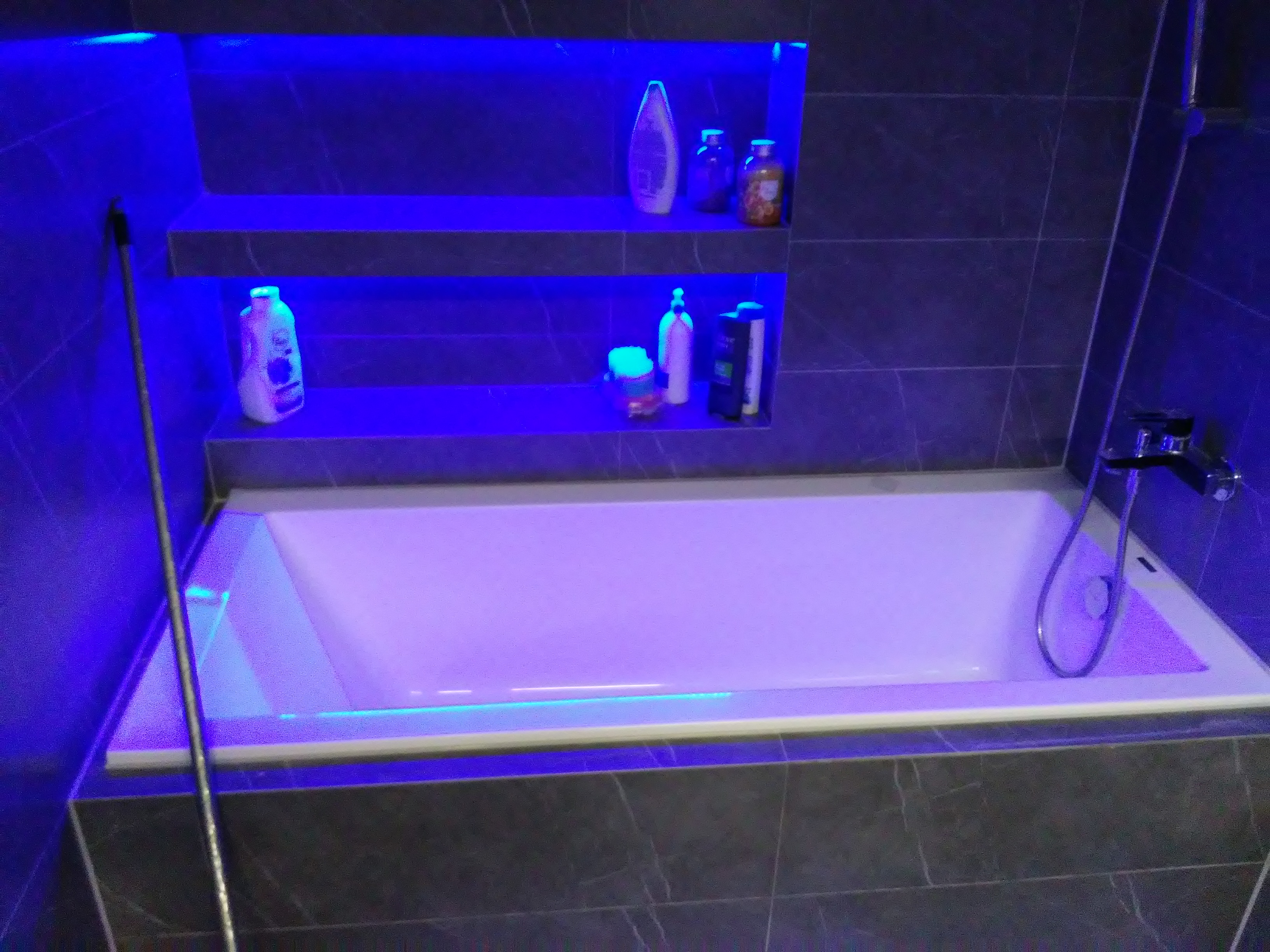 Wykończona łazienka z wanną, szarą glazurą na ścianach i półkami wnękowymi podświetlonymi niebieskim światłem LED, na których stoją kosmetyki.