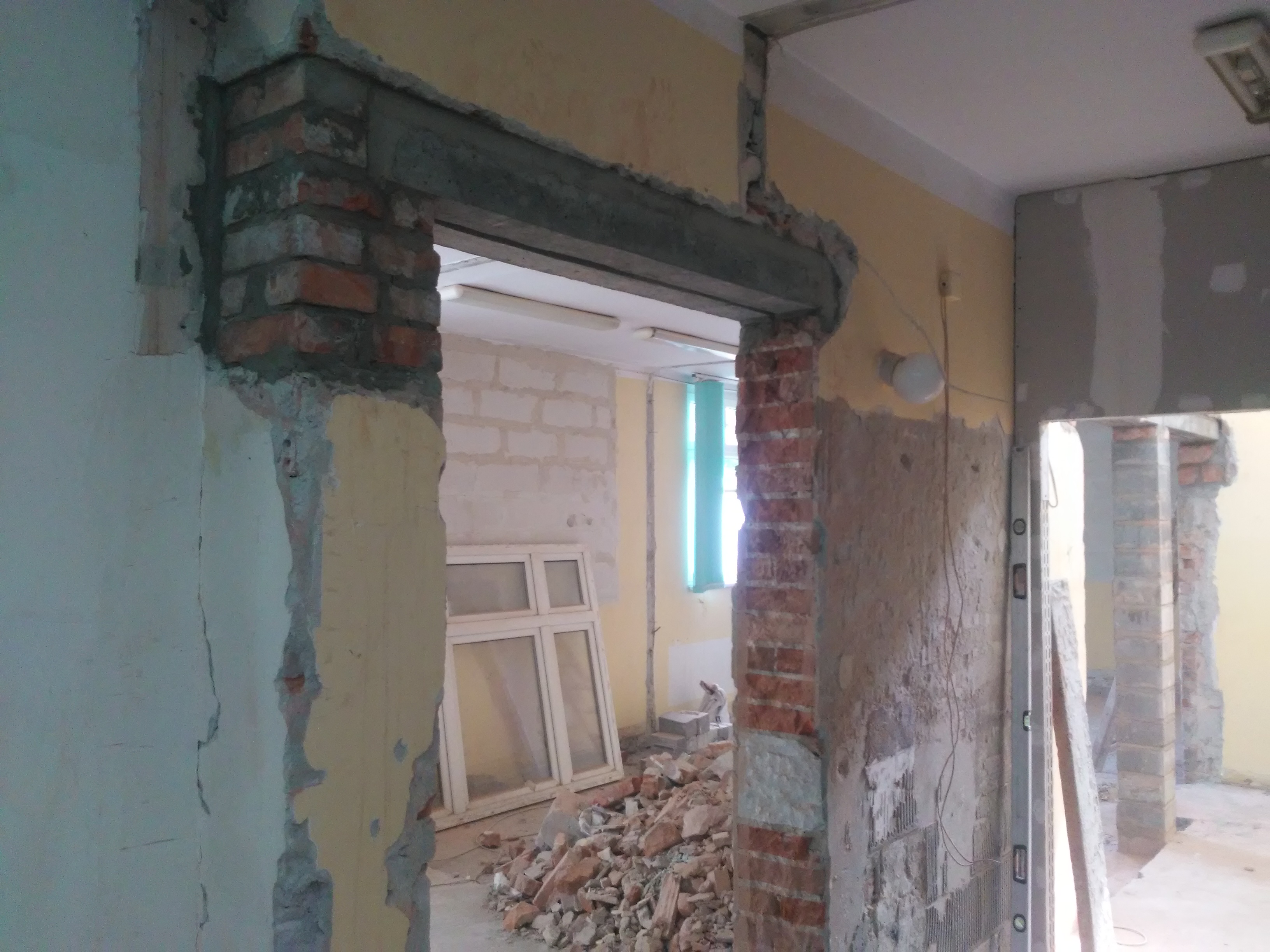 Otwór w ścianie z odsłoniętą cegłą, widoczne pozostałości gruzu i okno w tle, ślady po instalacjach elektrycznych na ścianie.