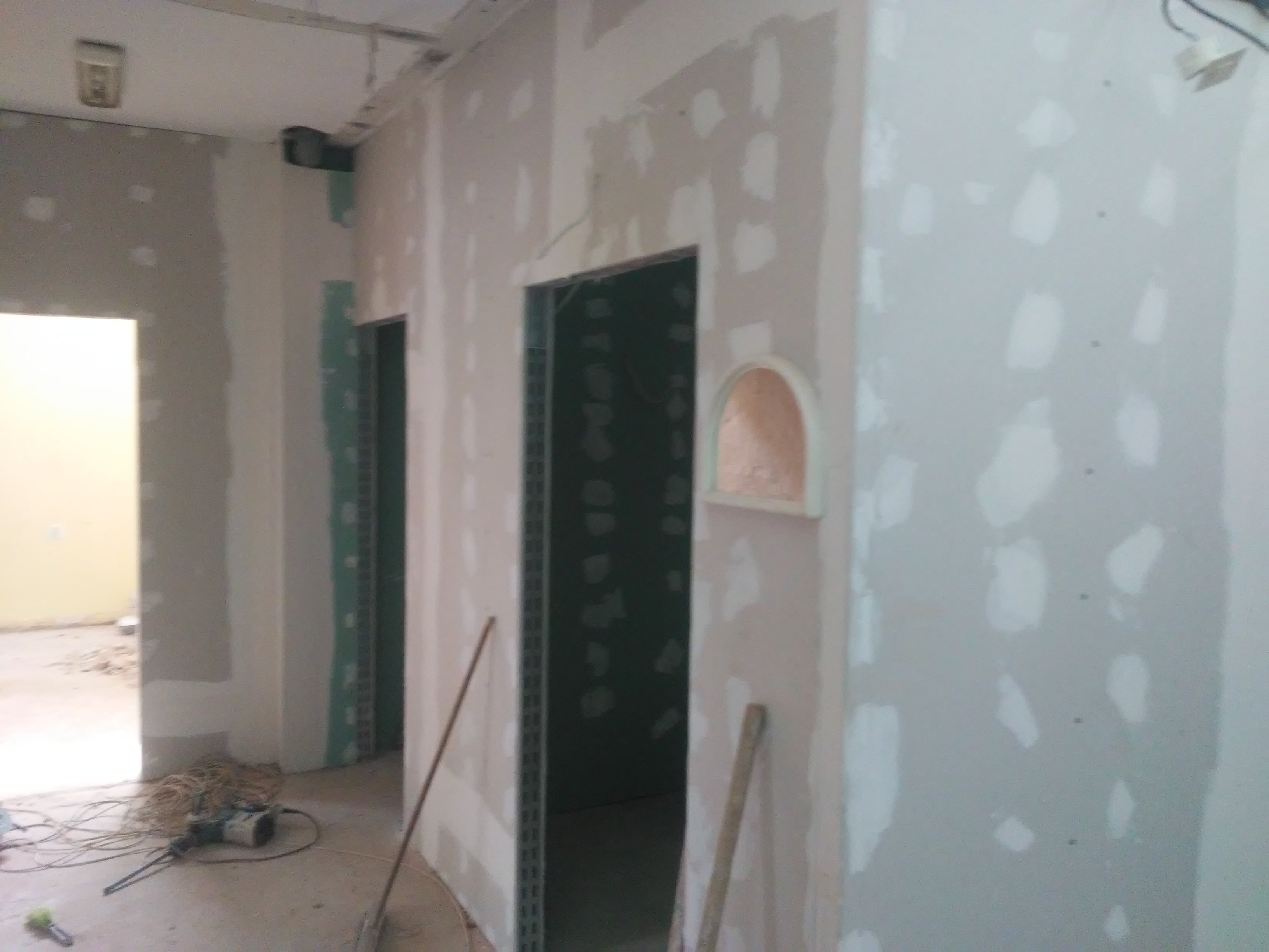 Wnętrze w trakcie prac wykończeniowych, widoczne ściany pokryte płytami gipsowo-kartonowymi z widocznymi łączeniami szpachlowanymi, otwory drzwiowe z zielonymi profilami, na podłodze narzędzia...