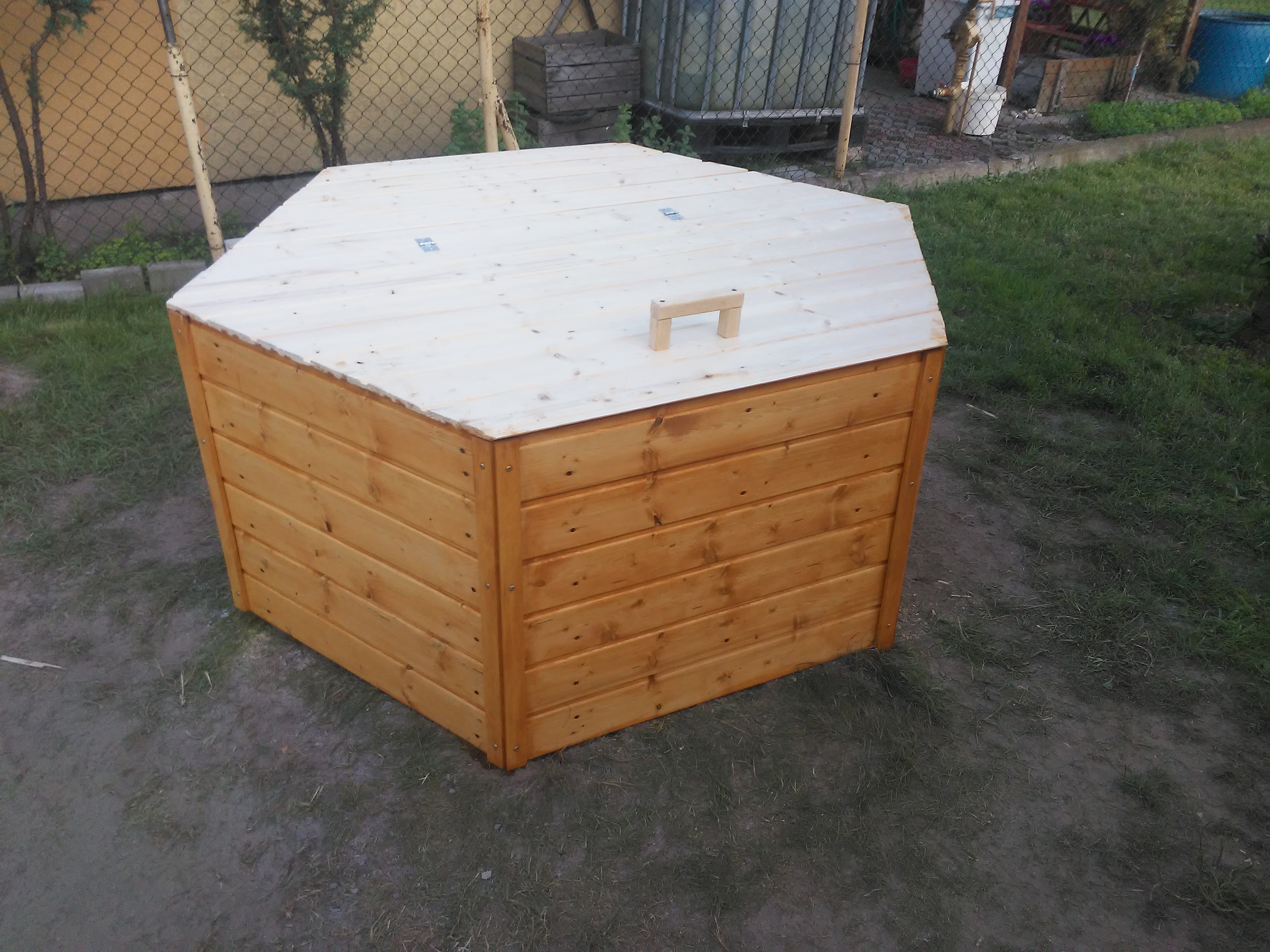 Drewniana skrzynia ogrodowa z jasną, sześciokątną pokrywą i uchwytem, stojąca na ziemi w ogrodzie.