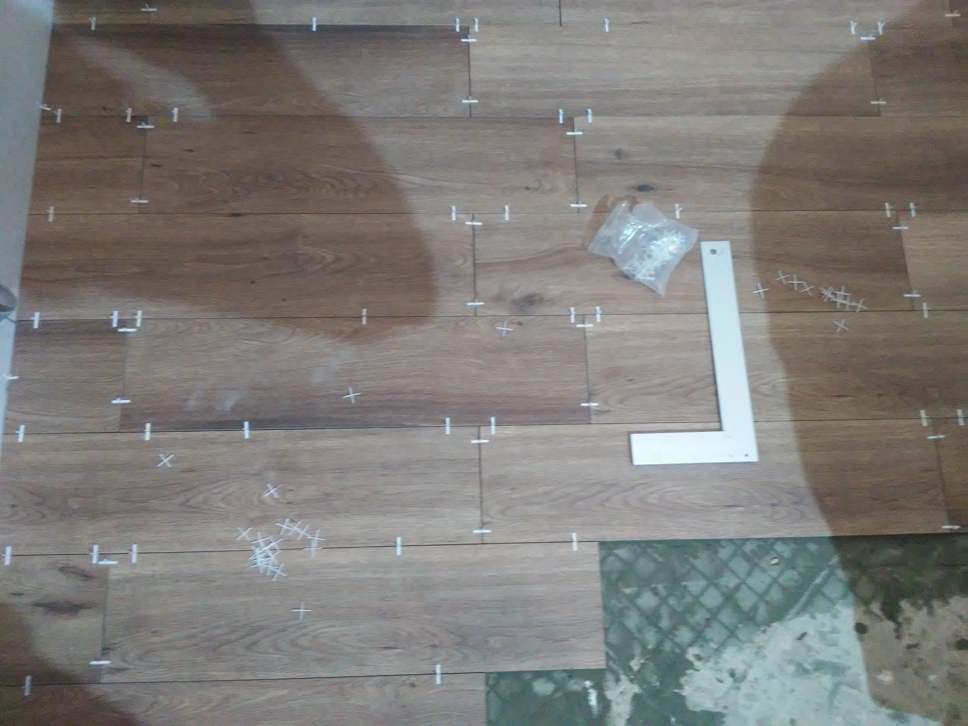 Układanie płytek podłogowych imitujących drewno, widoczne krzyżyki dystansowe w spoinach, poziomica kątowa, woreczek z krzyżykami i fragment surowego podłoża.