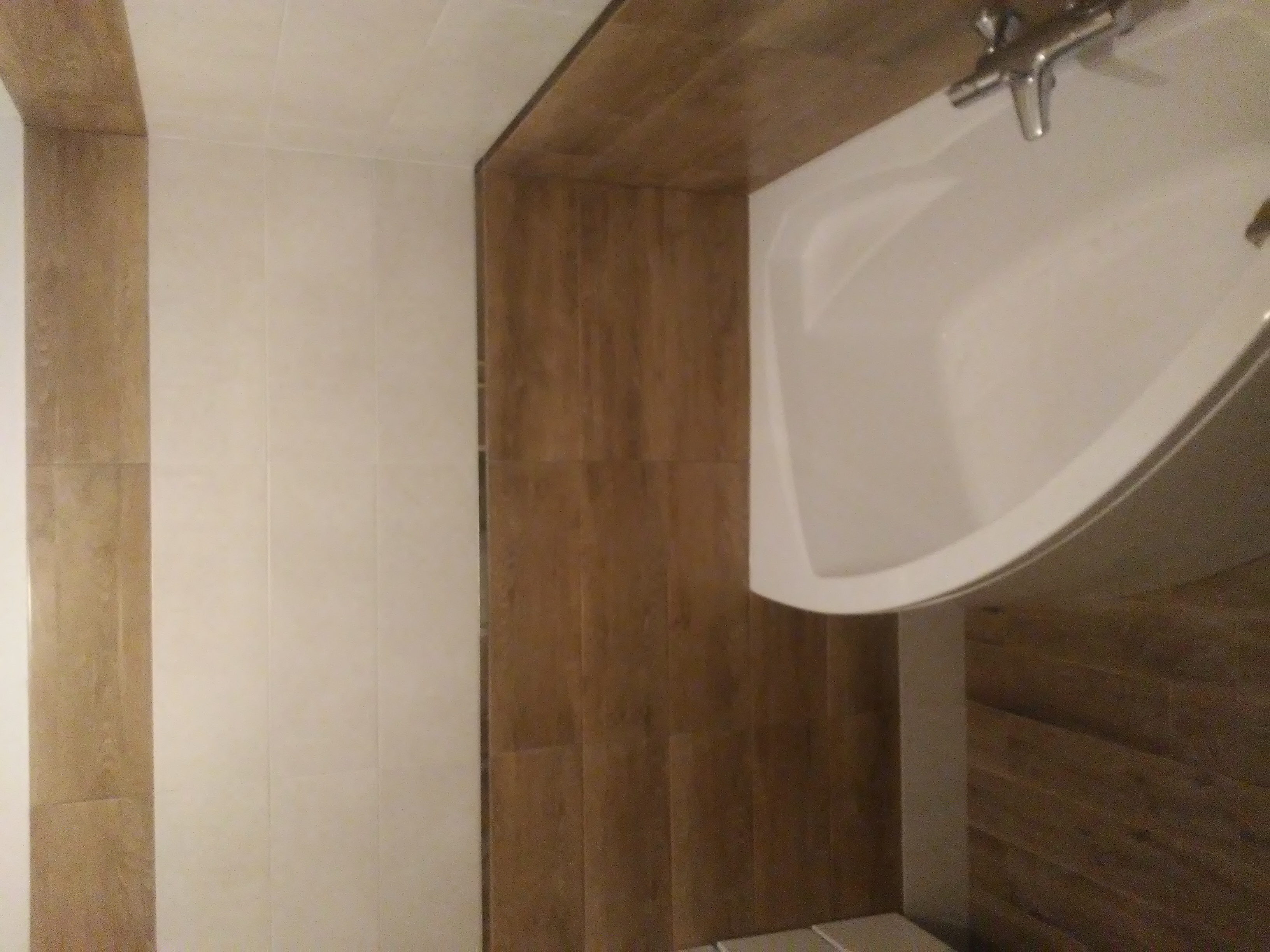 Wykończona łazienka z wanną narożną, jasnymi płytkami ściennymi i drewnopodobnymi płytkami na ścianie obok wanny, widoczna bateria wannowa.