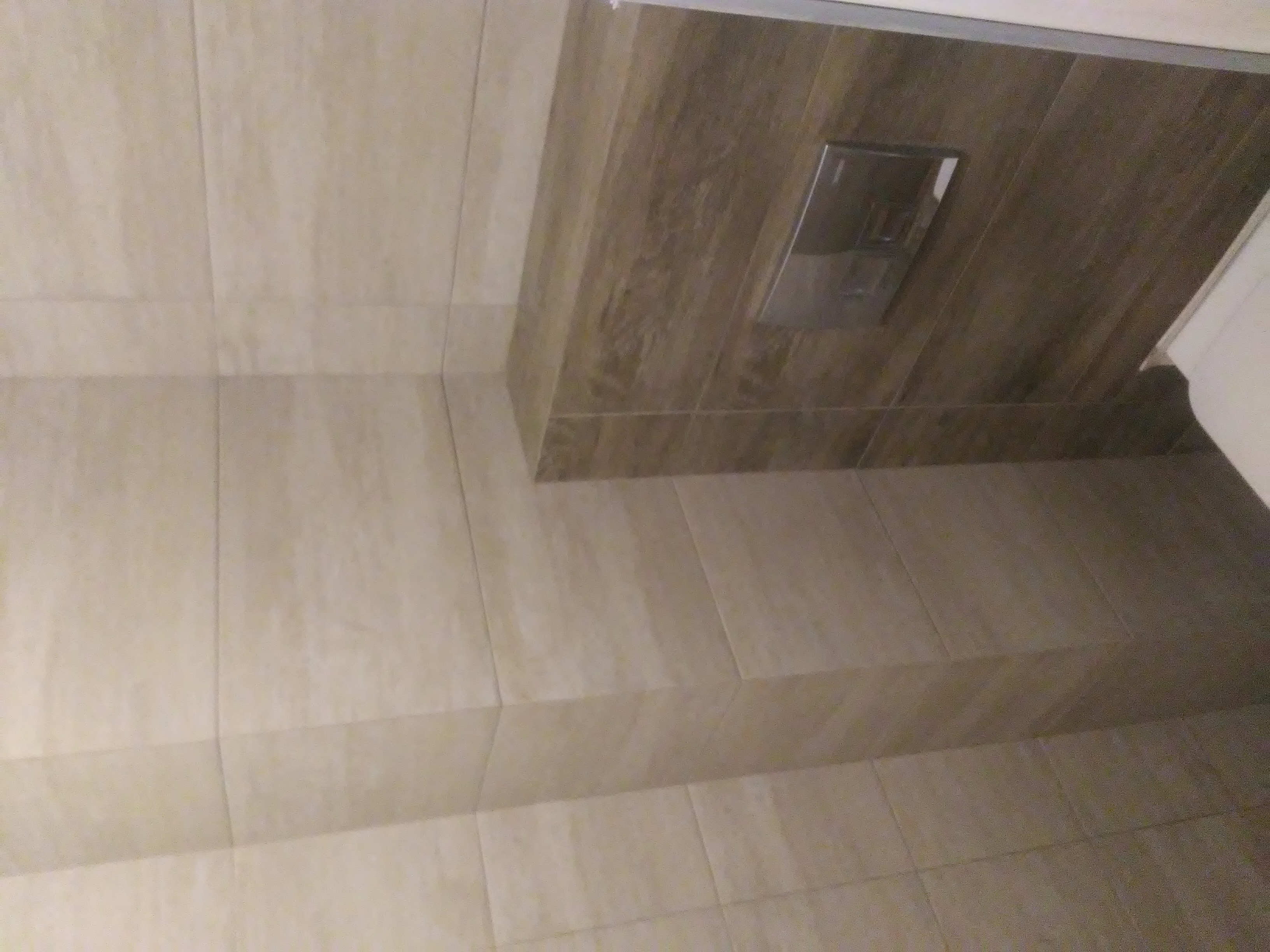 Beżowe płytki podłogowe i ścienne w łazience, z widocznym fragmentem toalety i przyciskiem spłuczki w obudowie z płytek imitujących drewno.