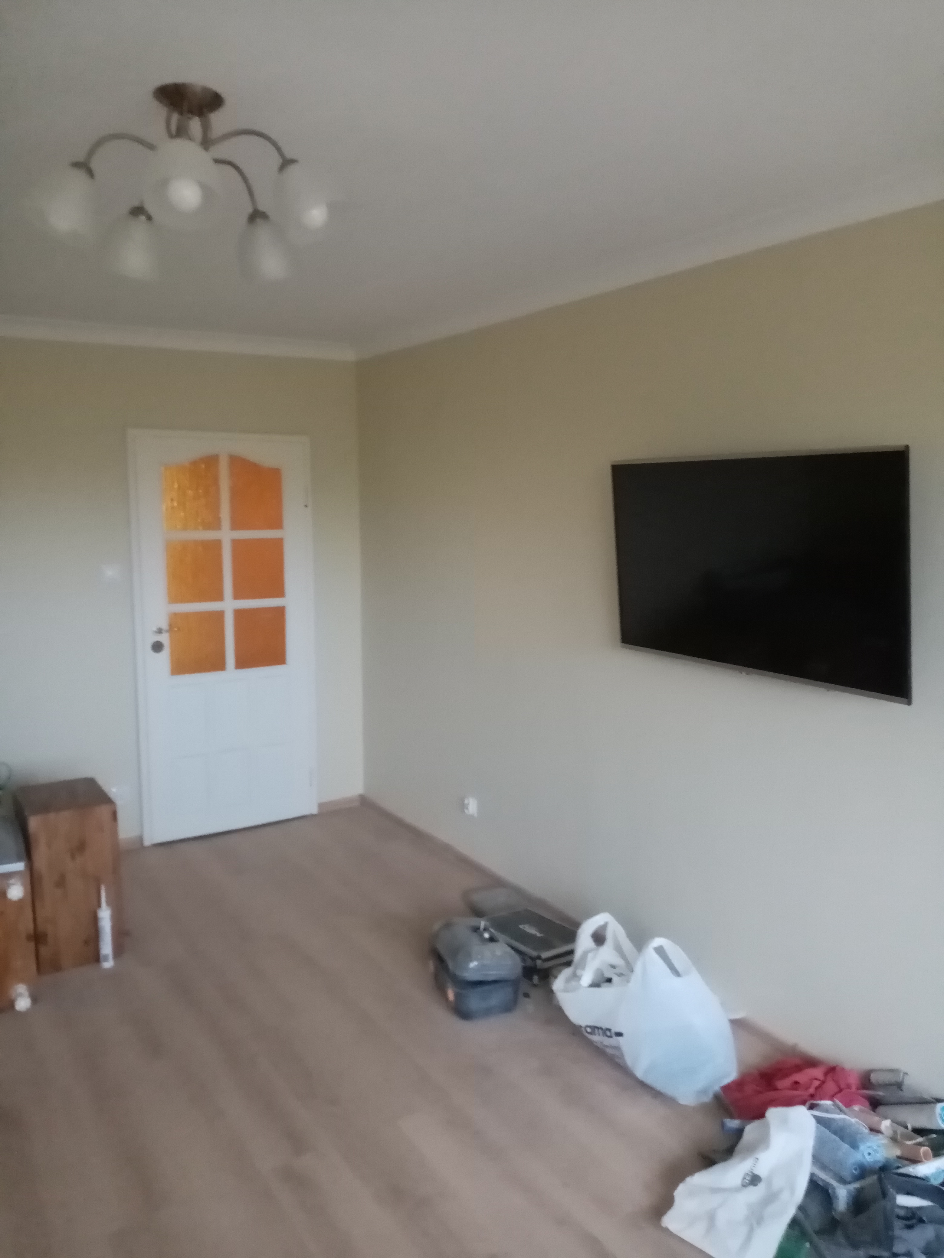 Wnętrze pokoju po remoncie z zamontowanym telewizorem na ścianie, widoczne narzędzia malarskie i białe torby na podłodze, białe drzwi z pomarańczowymi szybami w tle.