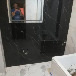 Czarne płytki imitujące marmur na ścianie łazienki z białymi żyłami, białe płytki podłogowe z szarymi wzorami, biała wanna oraz lustro z odbijającą się osobą robiącą zdjęcie.