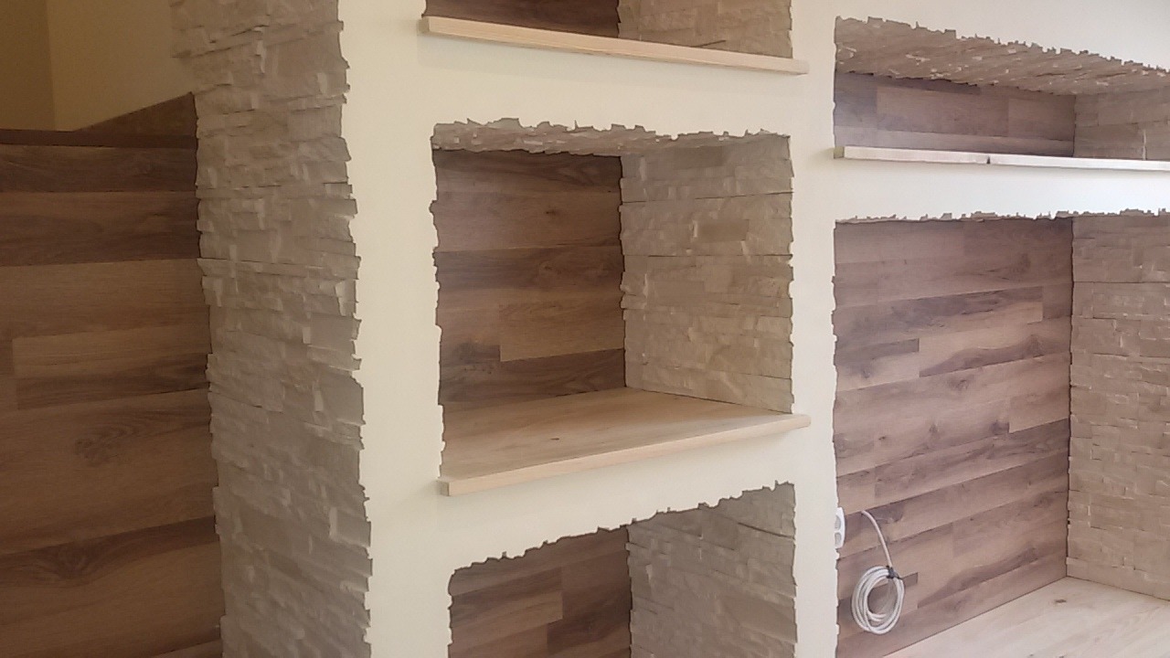 Ściana z wnękami wykończona kamieniem dekoracyjnym i panelami drewnopodobnymi, widoczne półki z surowego drewna, gniazdko elektryczne z przewodem.