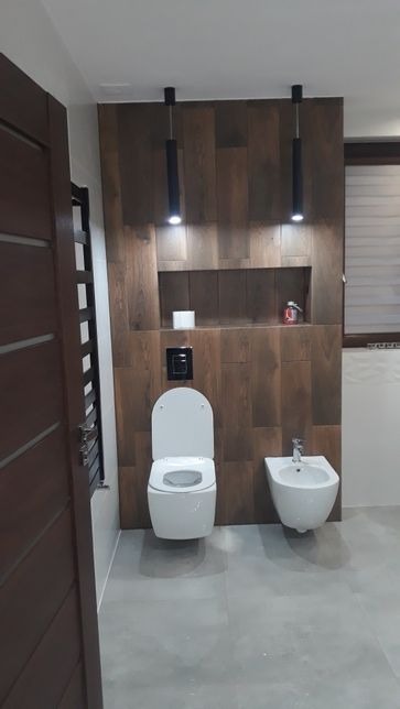 Nowoczesna łazienka z drewnopodobną okładziną ścienną, podwieszaną toaletą i bidetem, oświetlona wiszącymi lampami tubowymi, z szarą podłogą i widocznym fragmentem drewnianych drzwi.