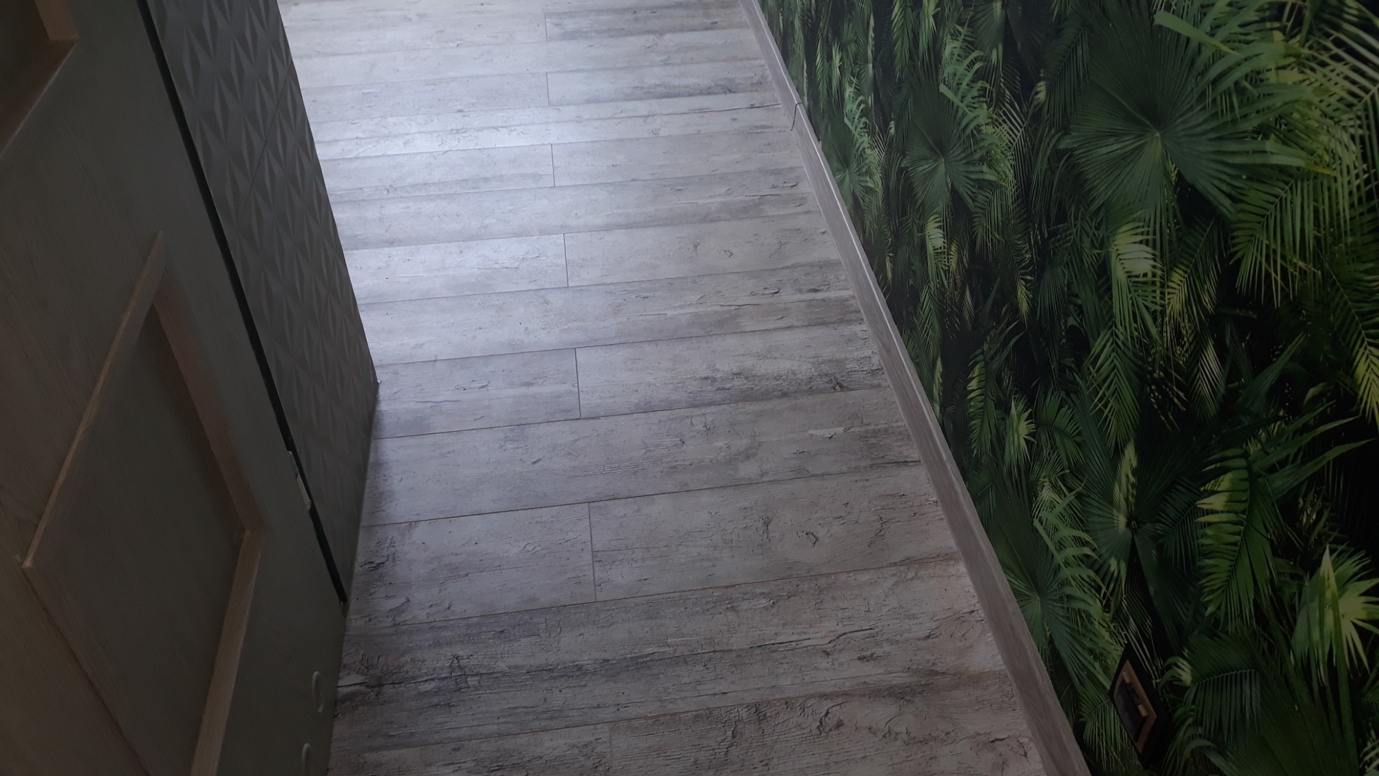 Szare panele podłogowe imitujące drewno, ułożone wzdłuż ściany z tapetą w motywie tropikalnych liści, widoczne fragmenty drzwi z geometrycznym wzorem.