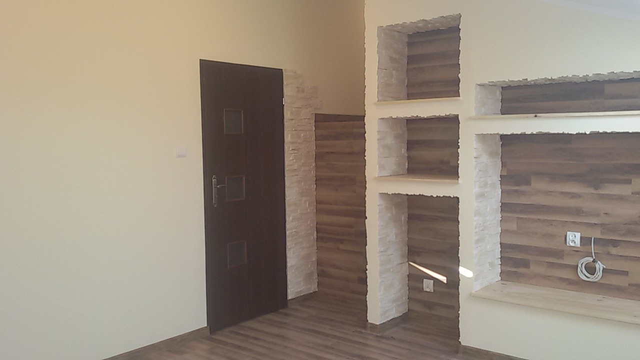 Ściana z wnękami wykończona imitacją kamienia i panelami drewnopodobnymi, obok ciemne drzwi wewnętrzne, widoczny fragment podłogi.