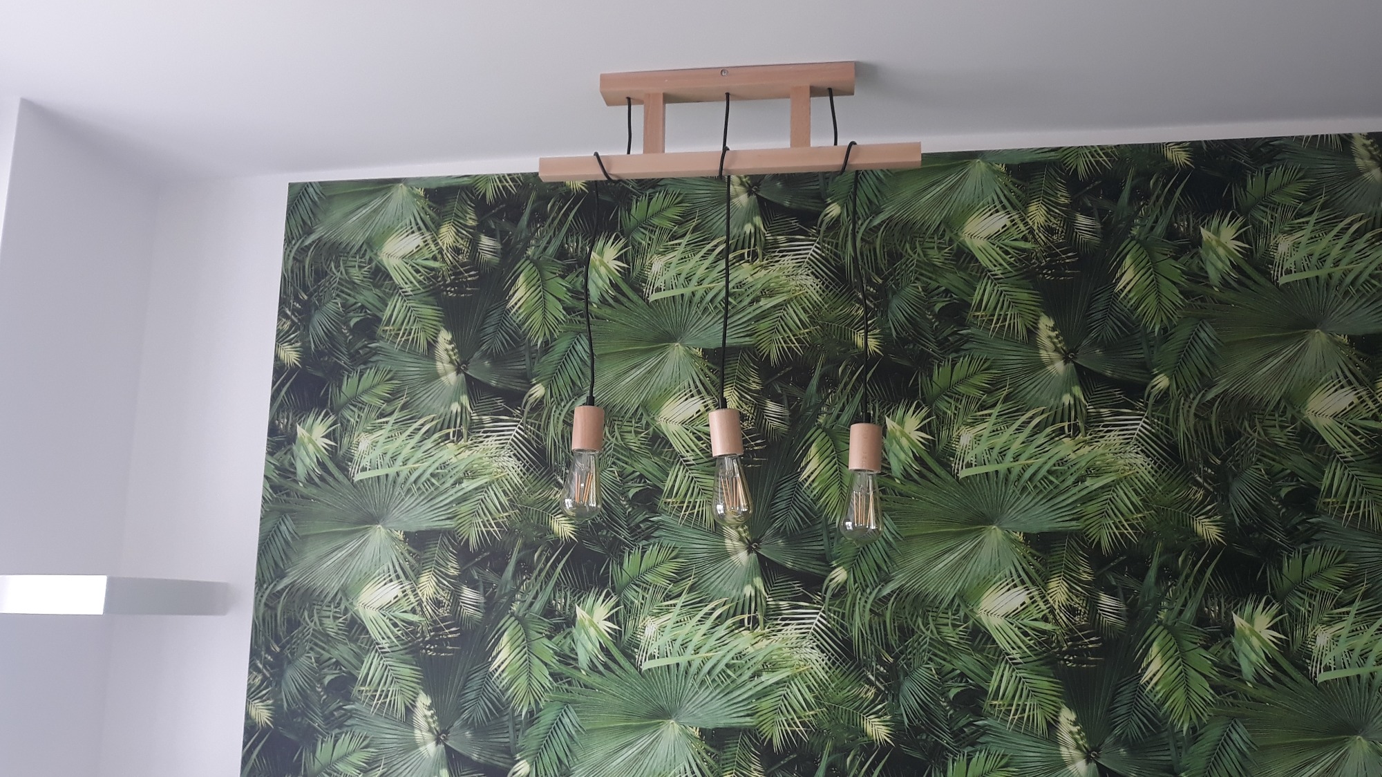 Lampa wisząca z trzema żarówkami Edisona na tle tapety z motywem tropikalnych liści palmy.