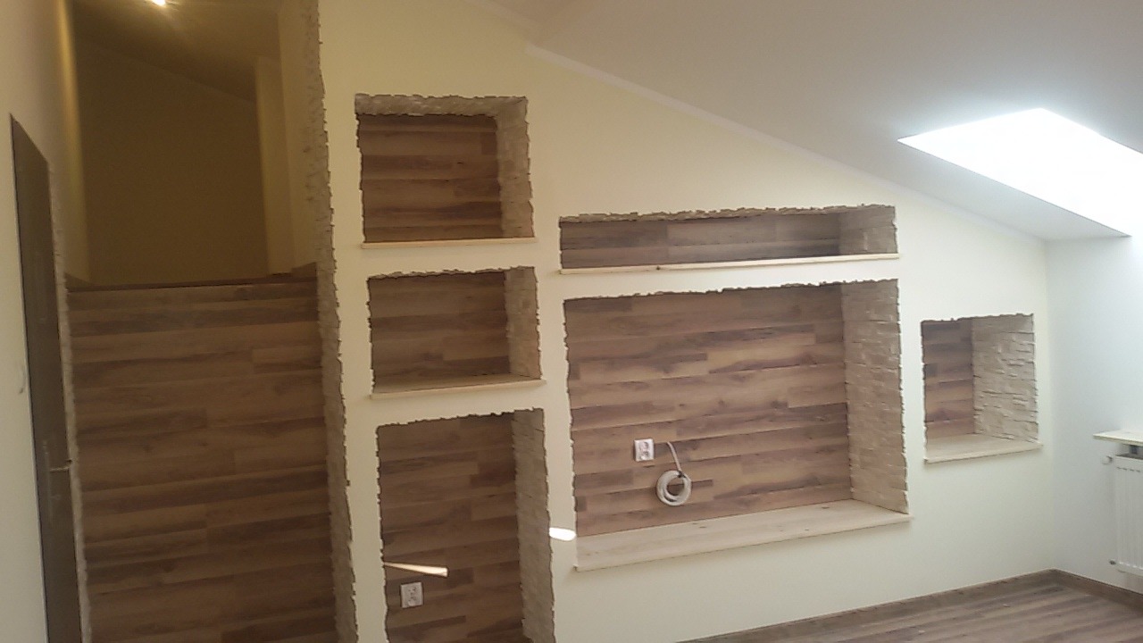 Ściana w trakcie prac wykończeniowych z wnękami obłożonymi panelami imitującymi drewno, widoczne gniazdka elektryczne i przewody, surowe krawędzie wnęk oczekujące na wykończenie.