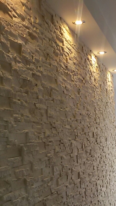 Ściana wykończona dekoracyjnym kamieniem w odcieniach beżu, oświetlona trzema okrągłymi lampami sufitowymi, widok pod kątem.
