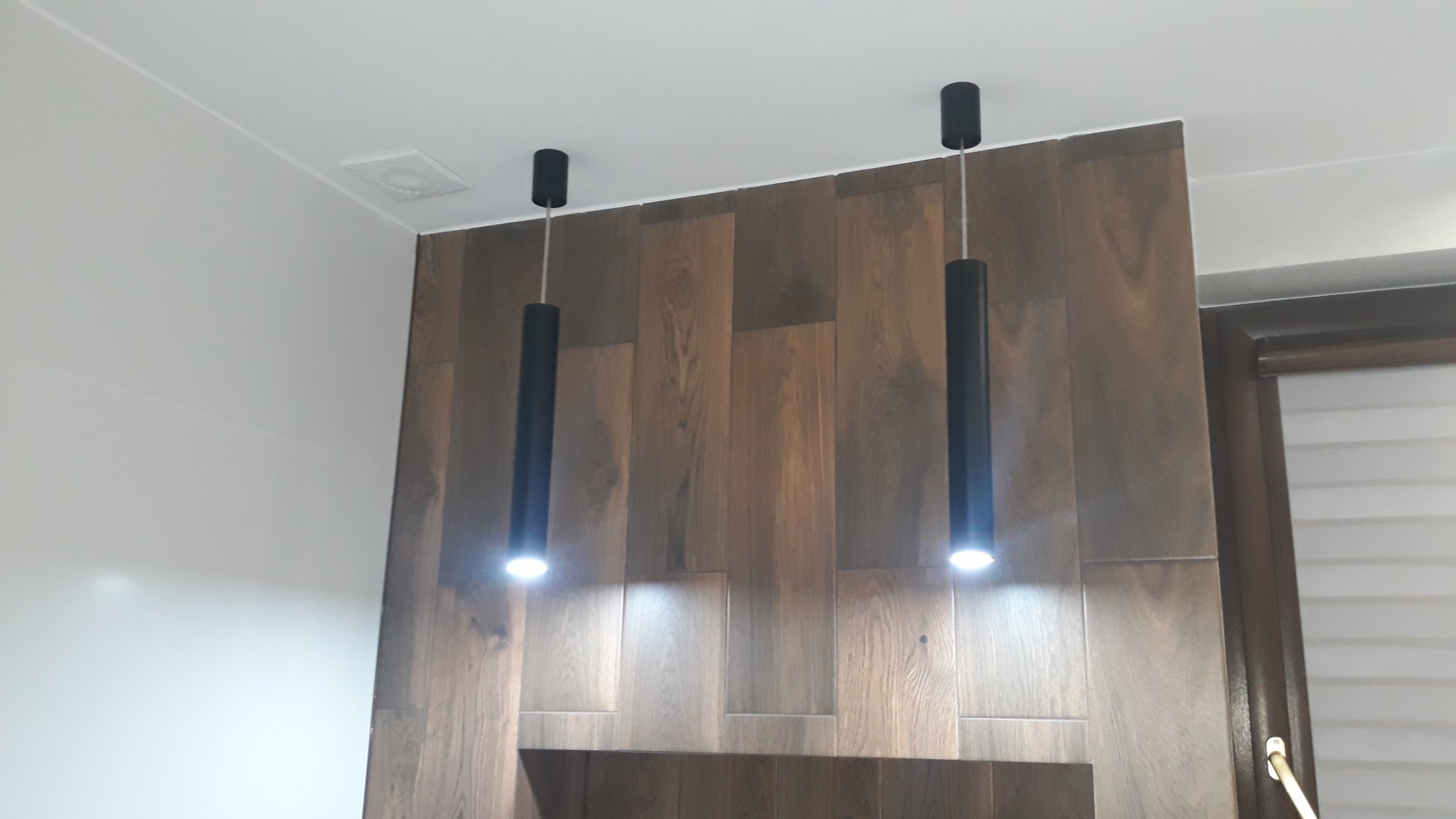 Dwa cylindryczne, czarne reflektory sufitowe LED oświetlają drewnianą ścianę z płytek imitujących drewno, widoczne także fragmenty sufitu i okna z żaluzjami.